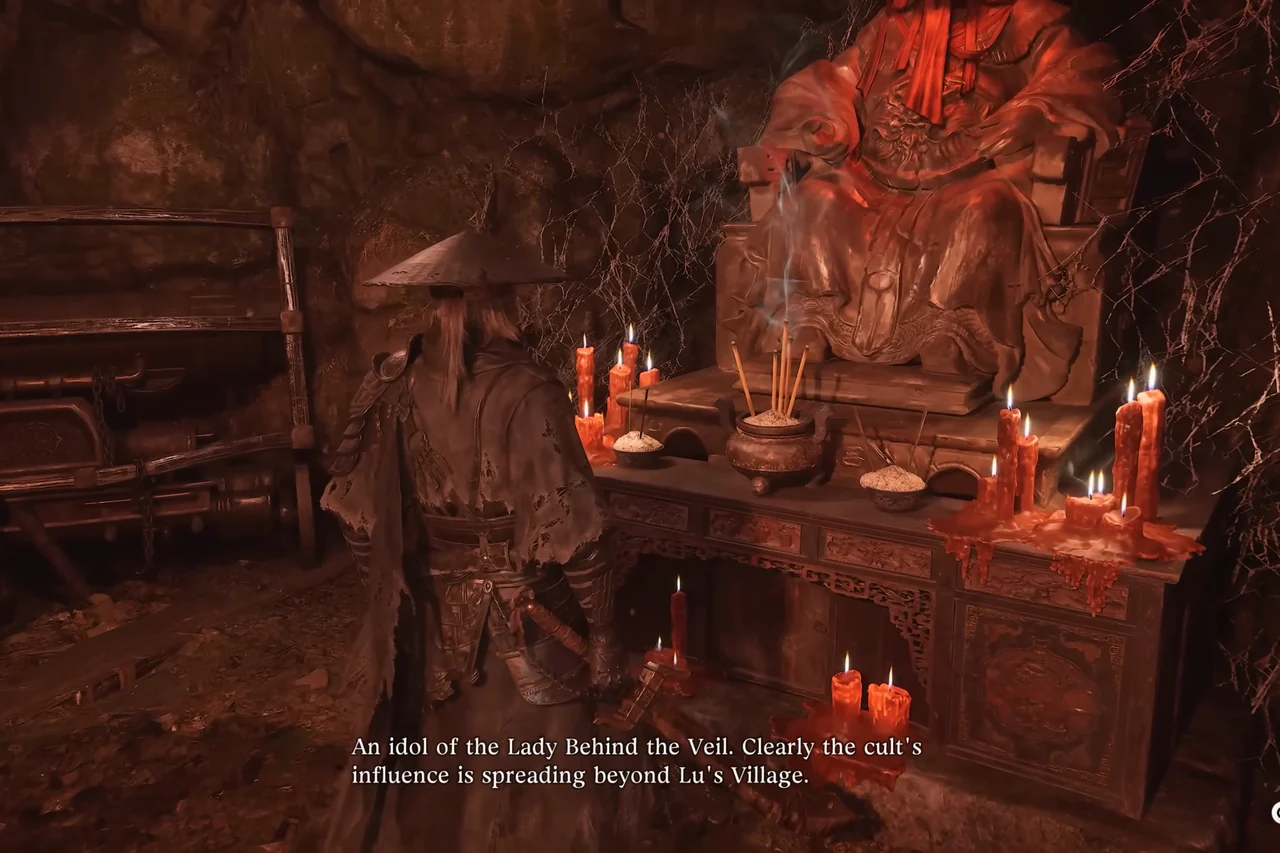 A seated idol among red candles in a cave shrine; a figure in a straw hat stands before it.