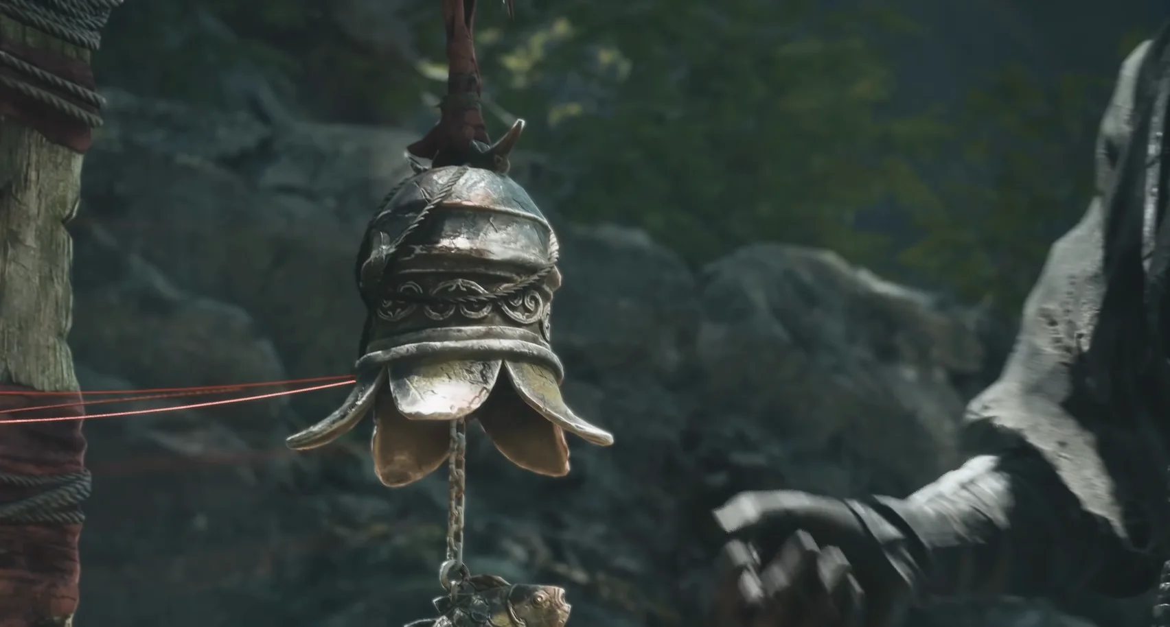 A bell-shaped object hangs from a post beside a tree; a gloved hand reaches in from the right.