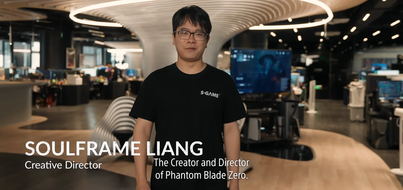 A man in glasses and a black T-shirt stands in an office; a name card at the lower left.